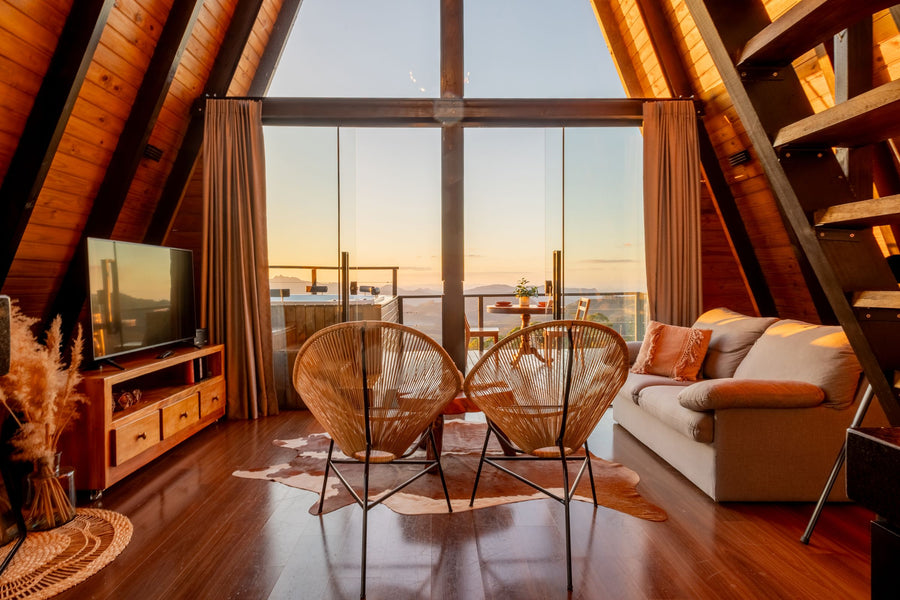 Wohnraum einer Ferienhütte im Chalet-Stil mit Holzgiebel, Rattansesseln und Panoramafenster mit Bergblick bei Sonnenuntergang